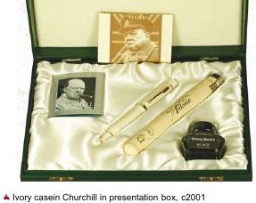 An open dark green presentation box lined in cream satin, holding a cream-coloured fountain pen and a small square bottle of black ink, with a portrait photograph set into the lid.