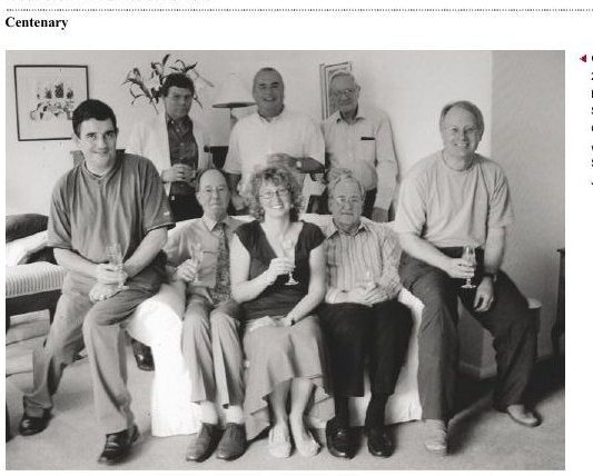 A colour photograph of eight people gathered in a sitting room, some seated on a sofa and some standing behind, several holding glasses.